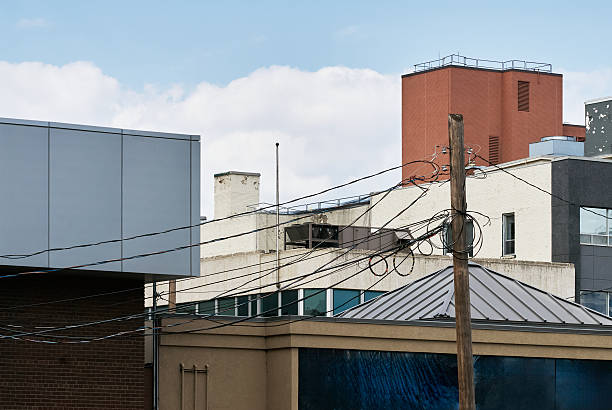 Cityscape view of multiple urban rooftops with mixed architectural styles, including brick and concrete buildings. A wooden utility pole with overhead power lines runs across the frame, creating a layered urban texture. The composition highlights infrastructure details, rooftop structures, and the contrast between modern and older architecture under a cloudy sky.