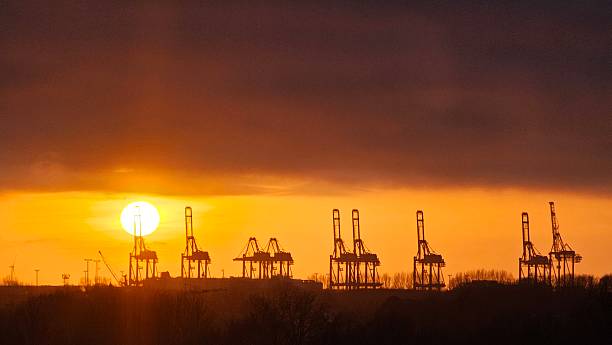 A line of massive harbor cranes is silhouetted against the vibrant orange and yellow sky of a sunset. The sky is partially covered by dark clouds, creating a dramatic contrast. The silhouettes of the cranes, trees, and other structures on the ground emphasize the industrial landscape of the port in Bremerhaven, Germany.