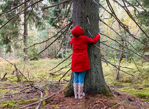 A middle-aged Caucasian woman in a red coat hugs a tree trunk.