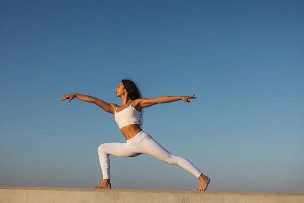 A woman dressed in white practicing yoga outdoors in a warrior pose. The scene captures a peaceful and focused moment set against a clear blue sky. Her pose demonstrates balance, flexibility, and strength, embodying a commitment to mental and physical wellness.