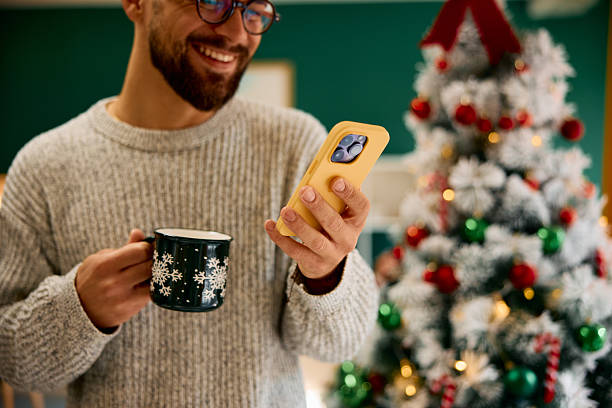istock Man smiling and using phone, holding a Christmas mug near Christmas tree 2235598397