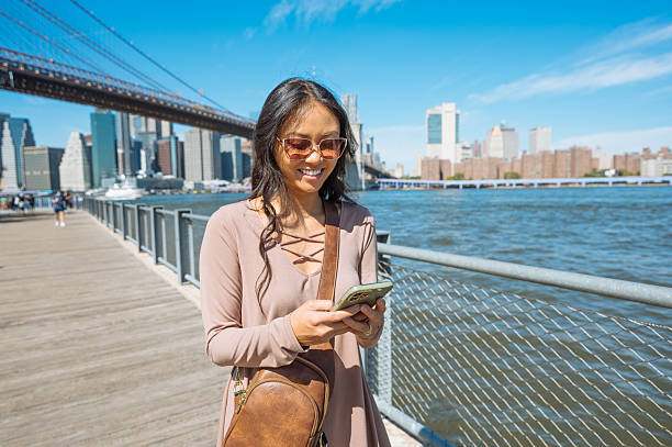 istock Woman smiling while using smartphone on a sunny day with iconic New York City skyline and bridge in the background 2235606054