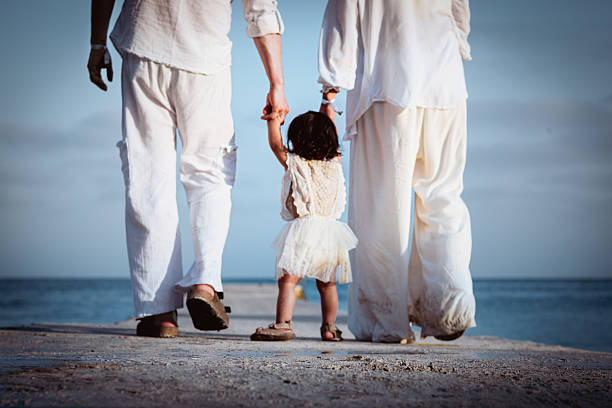 A family dressed in white walks along the coast at sunset. You can see the adults' feet and their daughter holding their hands, who is visible in full. They all have their backs turned.