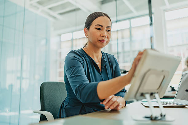 A mature asian woman is engaged in her work at a contemporary office. She is focused on a tablet while displaying a calm and confident demeanor, embodying professionalism and expertise.