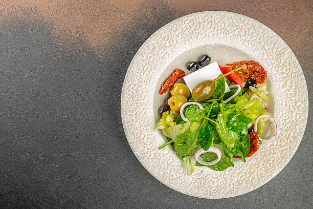Greek salad with feta cheese, olives, capers, sun-dried tomatoes, and fresh greens on a textured plate. copy space, top view