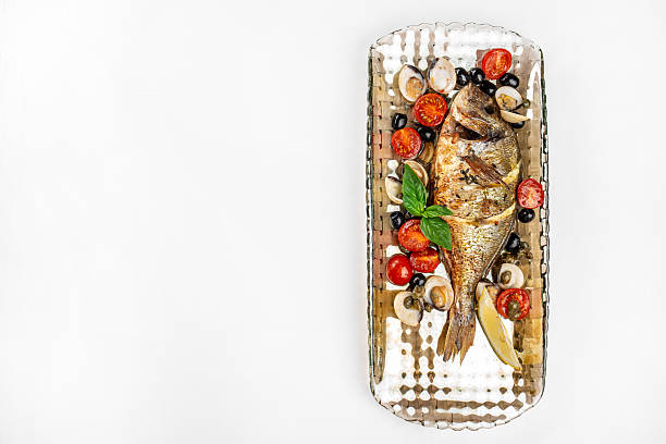 Grilled dorado fish with cherry tomatoes, olives, mushrooms, basil, and lemon on a metal tray. copy space, top view