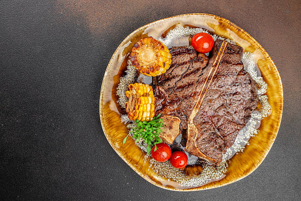 Grilled T-bone steak with corn on the cob, cherry tomatoes, and fresh herbs on a ceramic plate. copy space, top view.