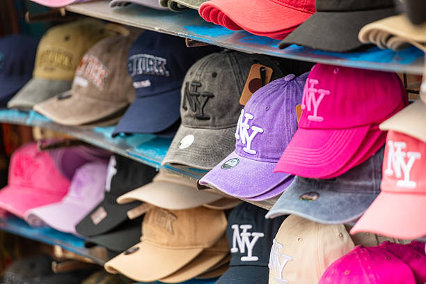 Close-up view of a colorful display of New York baseball caps arranged on shelves at a street vendor stall. The caps come in various styles and colors, including pink, purple, black, beige, and denim, many featuring the classic NY logo.