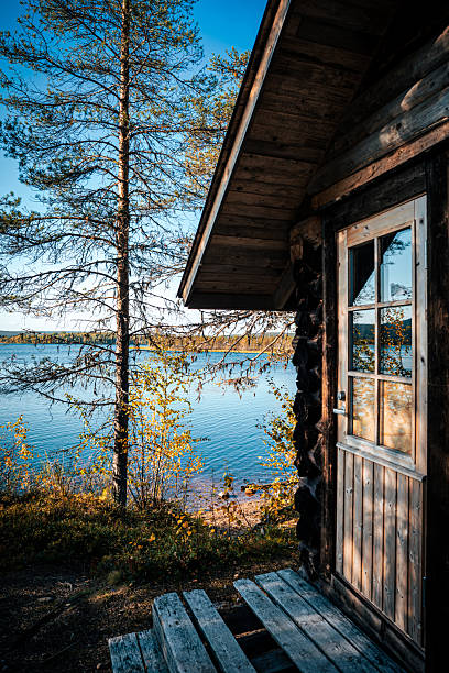 Cozy wooden sauna cabin on the shore of a lake in Lapland, Finland, with golden autumn trees reflected in the window. Calm water and warm sunlight create a peaceful Nordic wilderness atmosphere. Perfect for travel, lifestyle, and seasonal photography.