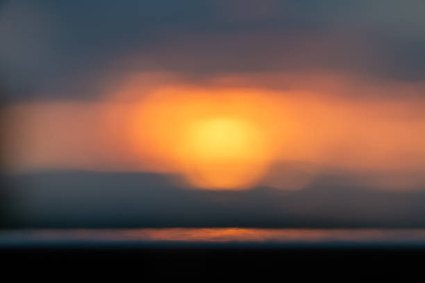 A dreamy, abstract sunset wallpaper with soft bokeh and warm central glow. The composition emphasizes color and light over detail, featuring a bright sun-like orb surrounded by smooth gradients of orange, yellow, and muted blues.