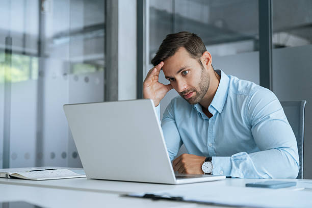 istock Confused young businessman looking disappointed at laptop pc computer screen having stress. European business man holding hand near temples, feeling stressed and tired sitting at workplace in office. 2235620327