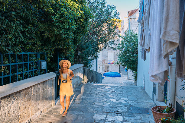 Young woman in yellow overalls and hat walking through a narrow stone alleyway with greenery. Female traveler with camera enjoying urban exploration. Solo adult on vacation in old town.