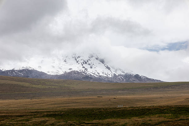 Snow-capped mountain rising above the high plateau, with heavy clouds covering its summit and open grassy plains stretching below.