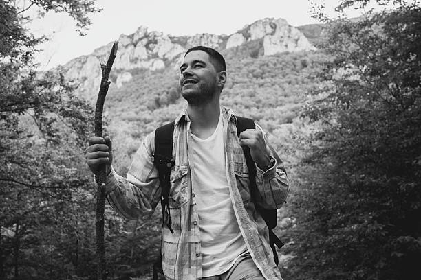 Black and white scene of a man hiking in the mountains. The individual is carrying a stick and backpack, expressing adventure and serenity surrounded by forest nature and rocky overviews.