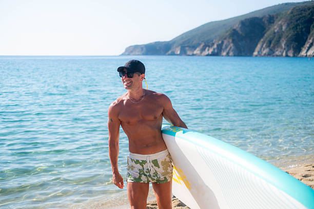 Muscular man is carrying his paddle board on a tropical beach, ready for a day of fun in the sun