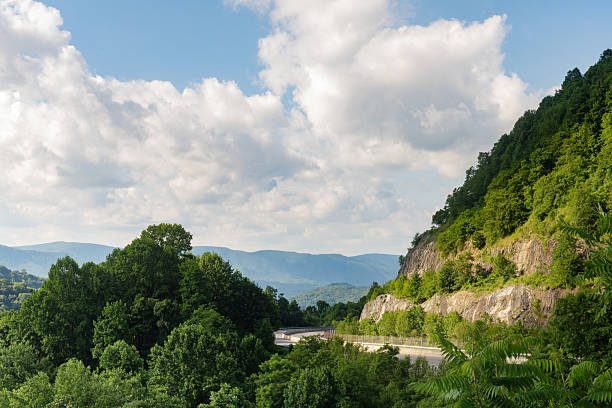 This is a photograph of a rural highway winding through lush trees by the Sams Gap Scenic Overlook in the Blue Ridge Mountains on a sunny day with clouds during summer in Flag Pond, Tennessee.
