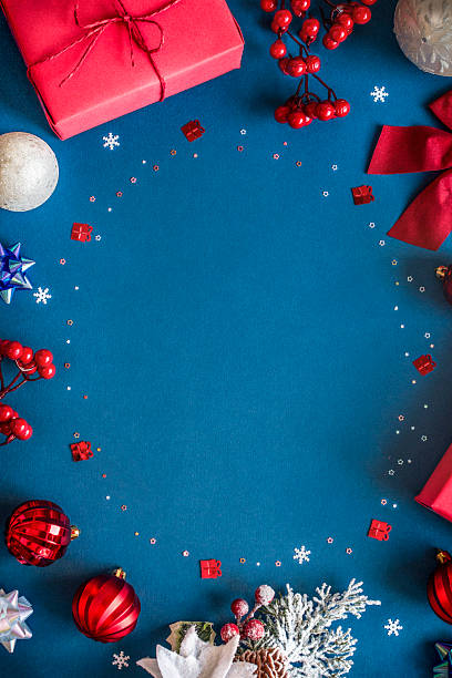 istock Vertical Christmas frame with red gift boxes, snowflakes, berries, frosted pine branches, trinkets, white and red baubles and ornaments on blue background with copy space in the middle. 2235645939
