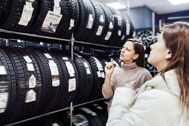 Two women engage in consultation at tire shop, discussing best options for seasonal tire purchases. One woman thoughtfully examines available tires displayed on shelves while seller provides guidance stock photo