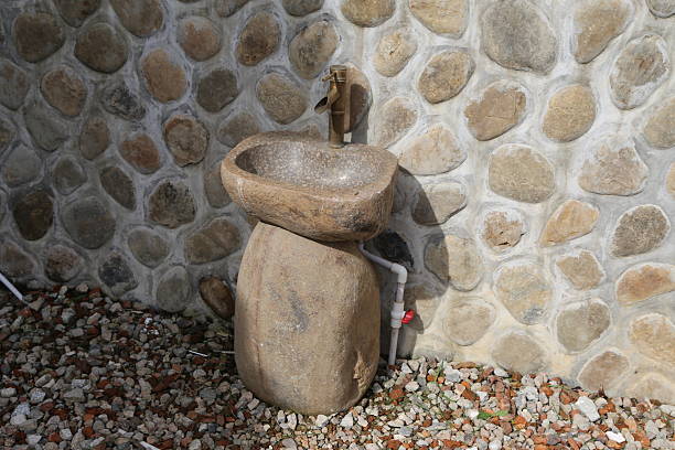 the stone sink is placed on the ground, with rough stones as its background wall and pebbles underneath it for decoration. the water pipe of an outdoor hand wash sits next to it.