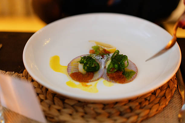 A close-up view of a beautifully plated, gourmet appetizer featuring two scallops served on their shells on a large white plate. The dish is meticulously garnished with microgreens, a vibrant orange sauce, and a lemon wedge.