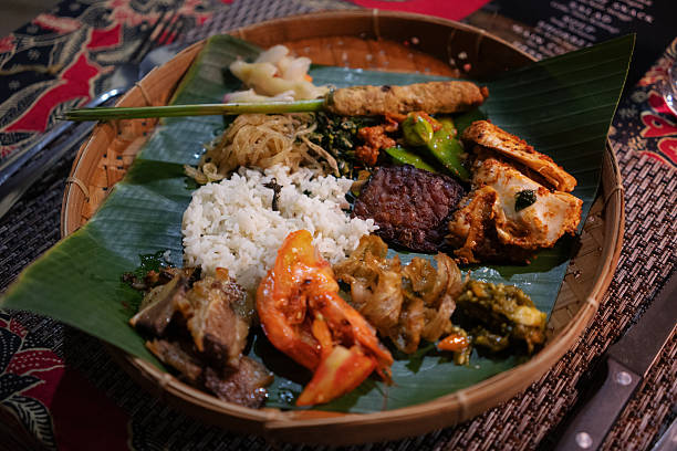 An overhead shot of a traditional Indonesian Nasi Campur dish served on a banana leaf placed on a woven bamboo tray. The meal consists of various components, including white rice, fried prawn, meat, and vegetables, all set on a table with a vibrant, red Indonesian batik tablecloth.