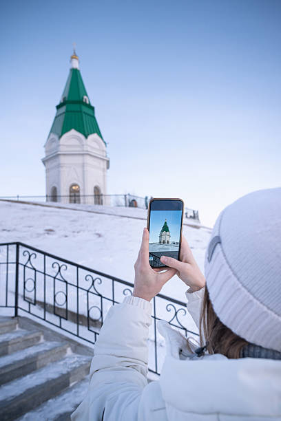 A woman takes pictures on her phone of the Paraskeva Friday Chapel, a Russian church on the site of the tower, the main attraction of Krasnoyarsk in Russia. Capturing the Enchanting Beauty of Winter A Historic Tower Majestically Draped in Snow stock photo