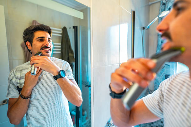 istock Young man shaving beard with electric razor in bathroom mirror 2235684605