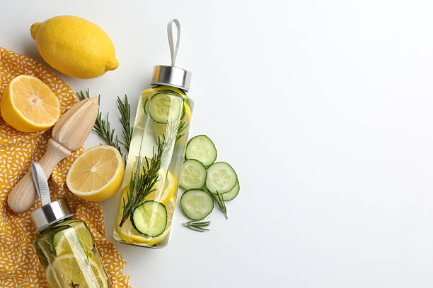 Lemon water in bottles, rosemary, fresh fruits, slices of cucumber and squeezer on white background, flat lay. Space for text