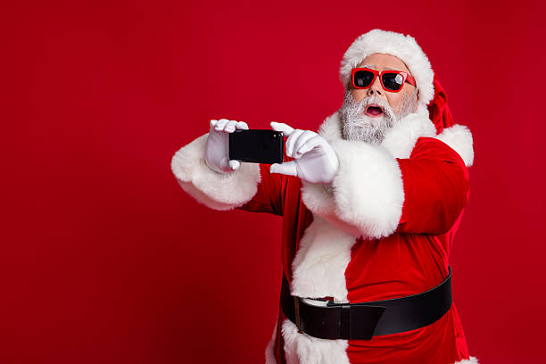 istock Portrait of attractive bearded amazed funny Santa using device taking making selfie vlog isolated over bright red color background 2235690899