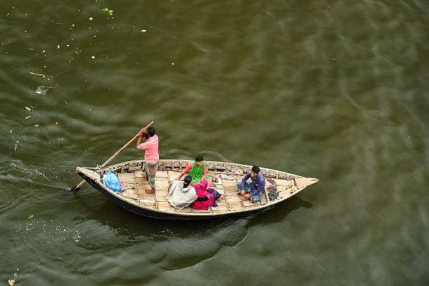 Dhaka, Bangladesh - 28 June, 2019: A group of individuals seated in a small wooden boat being paddled across a calm river, creating a serene and peaceful atmosphere.