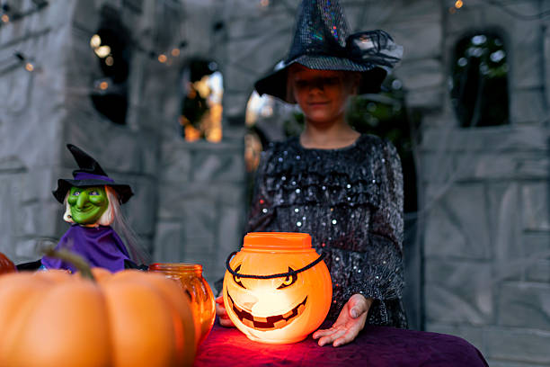 Child dressed as a witch standing behind a table with glowing jack-o’-lantern buckets and Halloween decorations. The spooky atmosphere is enhanced by pumpkins, a green witch figurine, and festive lights, creating a magical Halloween night scene.