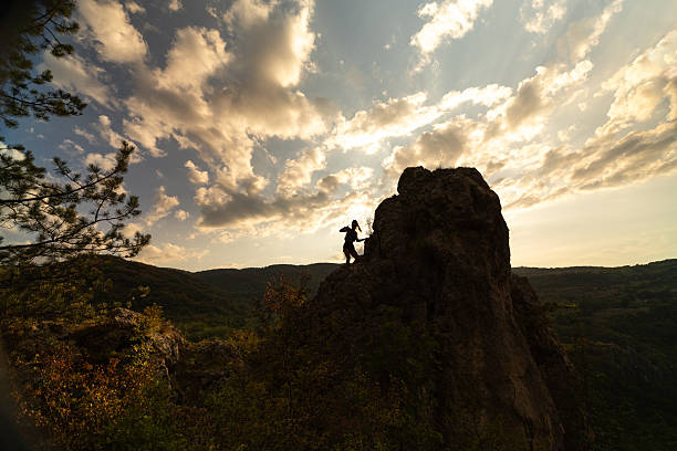 A silhouette of a hiker bravely ascending a rugged rock face, set against a scenic view of a dramatic sunset sky with scattered clouds, surrounded by a picturesque forested mountainous landscape.