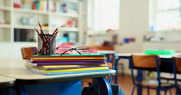Books, colors and glasses on desk in classroom for learning, education or knowledge with teaching. Spectacles, pencils and lesson at elementary school for assessment, test or project at academy.