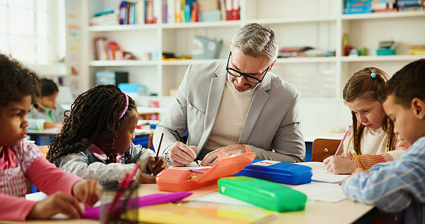 istock Education, learning or writing with students and teacher at desk in classroom for child development. Future, growth or notes with educator man teaching kids in elementary school class for lesson 2235704749