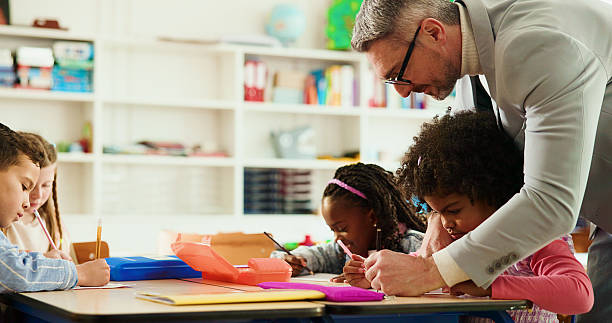 istock Education, teacher and man with children, writing or helping students with assignment or elementary. Knowledge, growth and development of kids in classroom, educator and teaching learner in school 2235704752