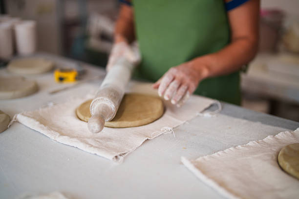 Hands of an artisan using a rolling pin to flatten clay on a worktable, with tools and ceramic pieces around. The scene highlights the preparation process in pottery making, suitable for themes such as craftsmanship, handmade ceramics, and artisan work.