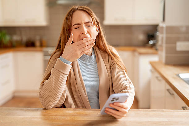 istock Woman yawning while using smartphone in cozy kitchen during morning routine with soft natural light filtering through the window 2235709531