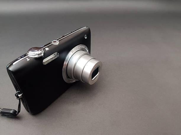 A sleek, black compact digital camera rests on a dark surface, illuminated by warm, atmospheric lighting. A laptop is partially visible in the background.