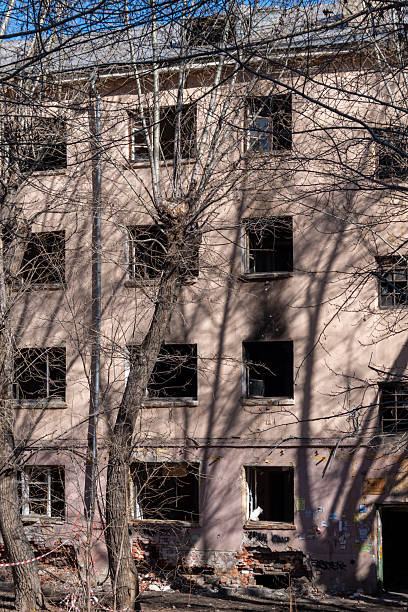 istock Old abandoned apartment building with shattered windows and damaged brickwork, surrounded by bare trees casting shadows on the wall 2235721893