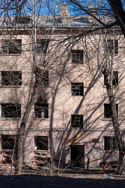 istock Old abandoned apartment building with shattered windows and damaged brickwork, surrounded by bare trees casting shadows on the wall 2235721894