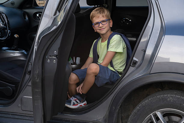 istock Boy with backpack sitting in car back seat looking at camera with smile. Safe transportation of children to school. A child goes to school by car 2235722120