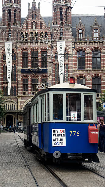 This editorial photo, taken on August 17, 2025, in Amsterdam, captures historic tram route No. 20 at Dam Square. The tram travels along cobblestone tracks in front of the De Bijenkorf department store, known for its Art Deco architecture.