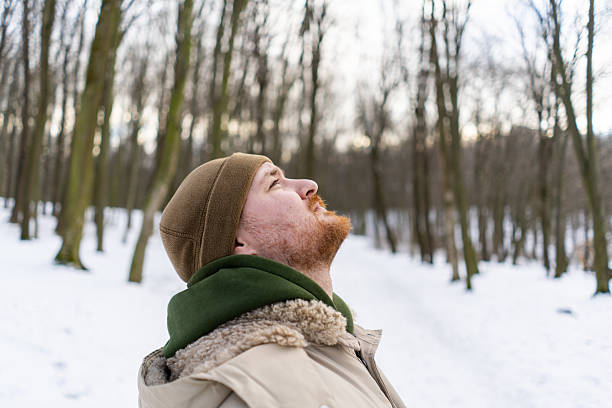 A man stands in a snowy forest. Trees surround him, and the winter air is crisp and cool. His warm clothing contrasts with the cold surroundings.