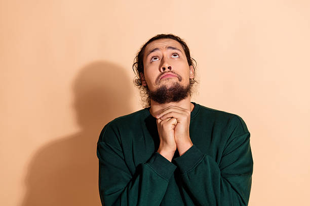 istock Young man making a pleading gesture with hands clasped and looking upwards over a beige background in casual attire 2235736611