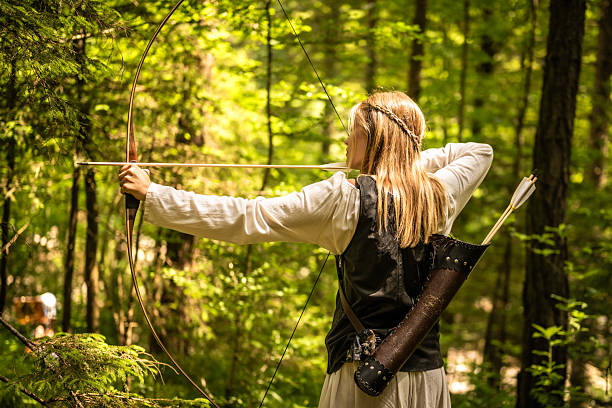 A woman in medieval attire is poised with a traditional wooden bow, aiming an arrow in a lush forest setting, captured with a american shot photo, her quiver, filled with arrows, is strapped to her back, evoking a sense of historical archery and outdoor sports training.