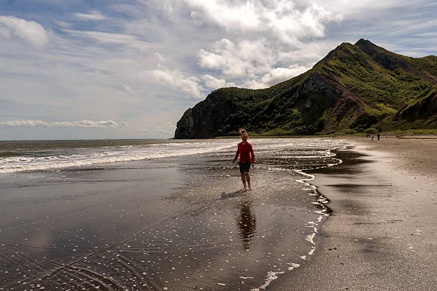 Girl in a red jacket walks along the foamy surf of a sandy beach, with dramatic mountain cliffs rising in the background. Tikhaya Bay in the Sea of ââOkhotsk, Sakhalin Island
