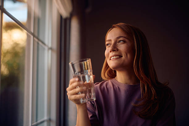 istock Smiling woman drinking water while standing by the window and looking through it. 2235740063