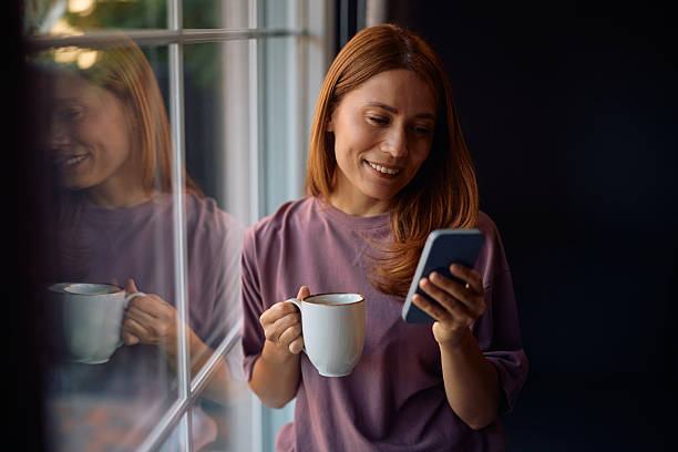 istock Happy woman with cup of coffee using cell phone by the window. 2235740100