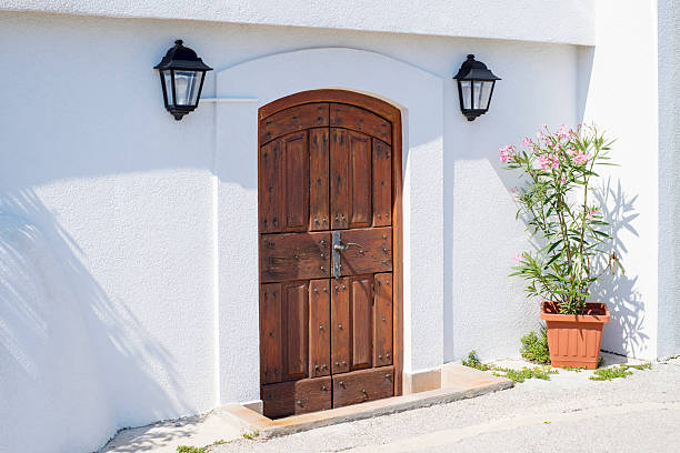 A rustic wooden door with black iron accents is set into a stone wall adorned with climbing plants. The door features an arched top and leads into a charming courtyard. The stone wall is made of irregular stones and is complemented by flower beds and a wooden pergola draped in greenery. This image evokes a sense of tranquility and rustic charm.