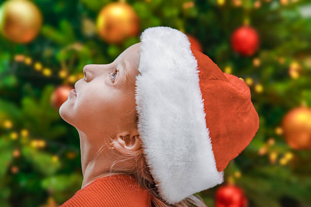 Festive portrait of a young child in a Santa hat gazing upward in wonder, with a decorated Christmas tree in the background. Concept of holiday magic, childhood dreams, joy, and festive celebration.